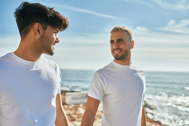 Νεαρό γκέι ζευγάρι χαμογελά ευτυχισμένος περπατώντας στην παραλία περιπάτους. - Φωτογραφία, εικόνα