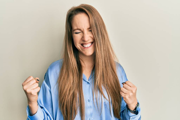 Jeune femme blonde portant une chemise bleue décontractée très heureuse et excitée faisant geste gagnant avec les bras levés, souriant et criant pour le succès. concept de célébration.  - Photo, image
