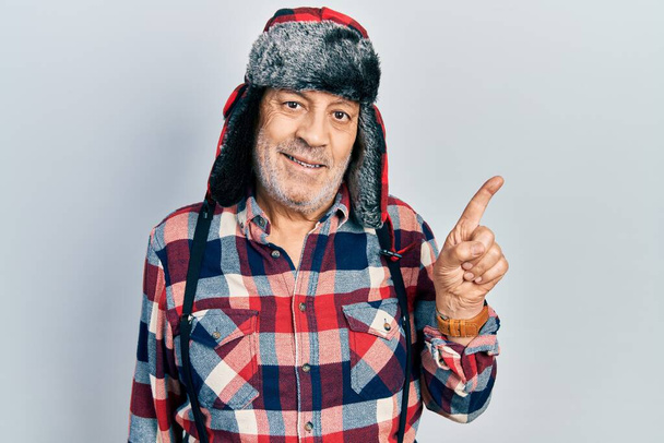 Bello uomo maturo che indossa cappello invernale con paraorecchie sorridente felice indicando con mano e dito di lato  - Foto, immagini
