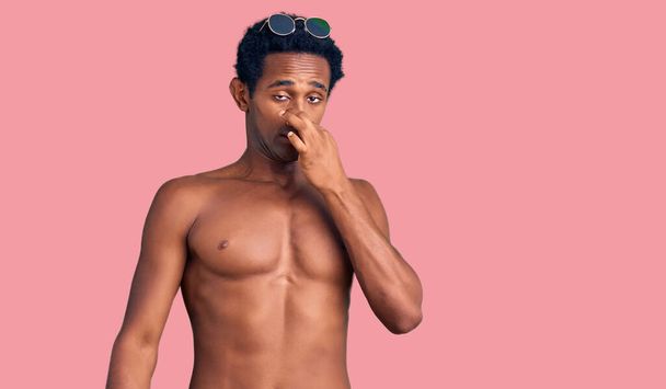 Afrikkalainen komea mies yllään uimapuku ja aurinkolasit haisee jotain haiseva ja vastenmielinen, sietämätön haju, pidätys hengitys sormet nenässä. paha haju  - Valokuva, kuva