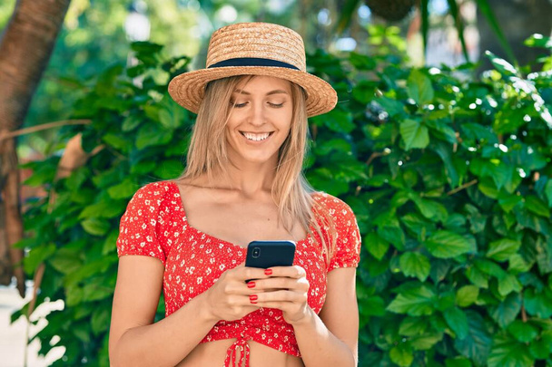 Νεαρή ξανθιά τουρίστρια που φοράει καλοκαιρινό στυλ χρησιμοποιώντας smartphone στο πάρκο. - Φωτογραφία, εικόνα