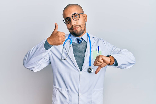 Latynoski dorosły mężczyzna w mundurze lekarza i stetoskopie, robiący kciuki w górę i w dół, niezgoda i wyrażenie zgody. szalony konflikt  - Zdjęcie, obraz