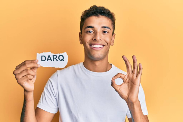 Junger gutaussehender afrikanisch-amerikanischer Mann, der Darq-Technologien in der Hand hält und mit den Fingern Okay-Zeichen macht, freundlich lächelnd, hervorragendes Symbol gestikulierend  - Foto, Bild