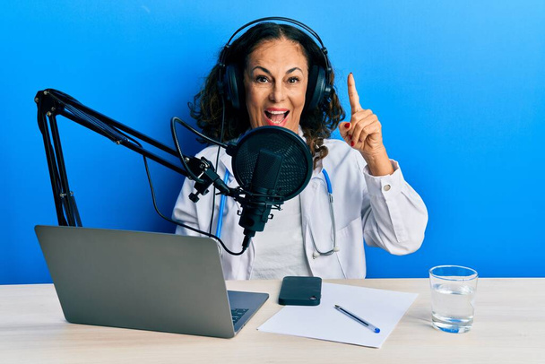 Mooie middelbare leeftijd dokter vrouw werkt bij radio studio wijzend vinger omhoog met een succesvol idee. opgewonden en gelukkig. Nummer een.  - Foto, afbeelding