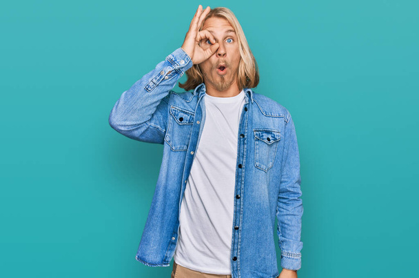 Caucasian man with blond long hair wearing casual denim jacket doing ok gesture shocked with surprised face, eye looking through fingers. unbelieving expression.  - Photo, Image