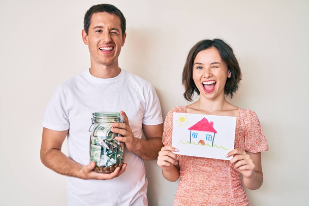 Hermosa pareja sosteniendo el dibujo de la casa y frasco con dólares guiñando el ojo mirando a la cámara con expresión sexy, cara alegre y feliz.  - Foto, imagen