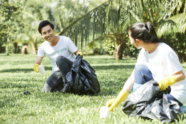 Happy young Asian couple in volunteer t-shirts collecting garbage in local park - Fotoğraf, Görsel