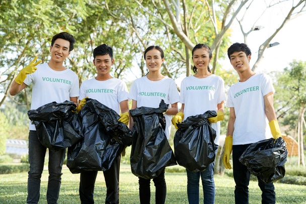 Giovani uomini e donne sorridenti in magliette volontarie in piedi nel parco cittadino con sacchi della spazzatura dopo aver raccolto spazzatura - Foto, immagini