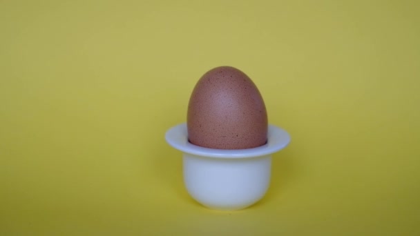 Ruka odstraňuje přírodní hnědé vejce z jednoduchého bílého vaječného šálku na žlutém pozadí. Snídaně. Minimalistický přírodní velikonoční motiv. - Záběry, video