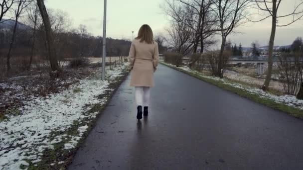 Dívka v zimním kabátě a bílé kalhoty kráčející po venkovské silnici během zatažené oblačnosti v zimě. V uličce se hromadí sníh - Záběry, video