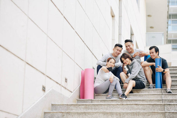 Grupo de jóvenes vietnamitas felices con colchonetas de yoga sentados en las escaleras y tomando selfie después de entrenar juntos - Foto, Imagen