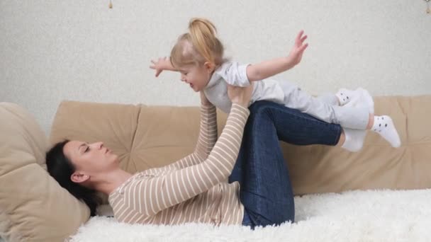 Happy beautiful family healthy young mom lifting cute little child daughter up playing plane on sofa at home, funny kid girl flying in mothers arms practice acro pair yoga bonding having fun together - Footage, Video
