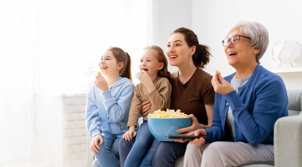 Feliz familia viendo televisión, películas con palomitas de maíz en casa. Madre, hijas y abuela pasando tiempo juntos. - Foto, imagen