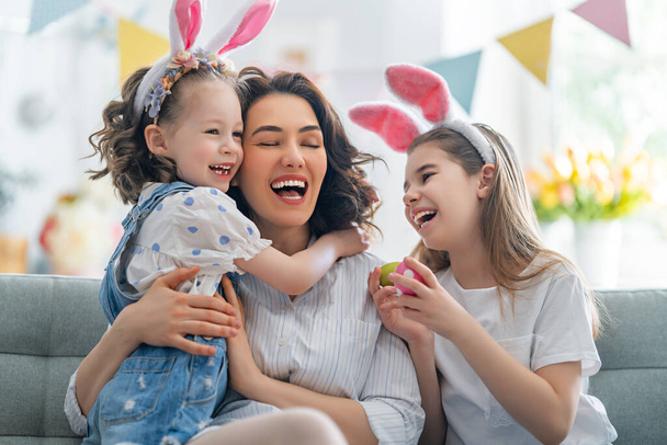 Happy holiday! Mother and her daughters with painting eggs. Family celebrating Easter. Cute little children girls are wearing bunny ears.  - Foto, Bild