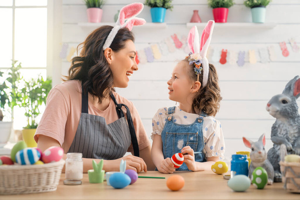 Mother and her daughter painting eggs. Happy family preparing for Easter. Cute little child girl wearing bunny ears. - 写真・画像