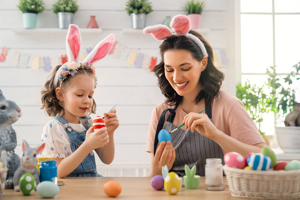 Mother and her daughter painting eggs. Happy family preparing for Easter. Cute little child girl wearing bunny ears. - Φωτογραφία, εικόνα