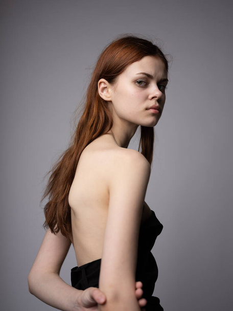 Beautiful woman with red hair in a black dress on a gray background bared shoulders - Foto, imagen