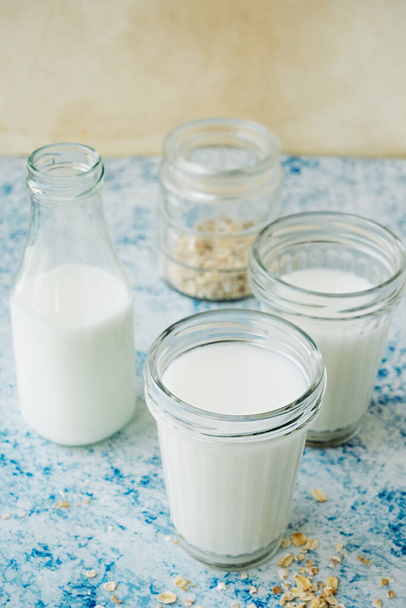 három csésze tej és üveg ital, szétszórt zabkása a kék asztalon. minimalizmus trendi élelmiszer-tartalom. növényi alapú alternatív tejfajta. zab tej készítésének folyamata otthon, szelektív fókusz - Fotó, kép