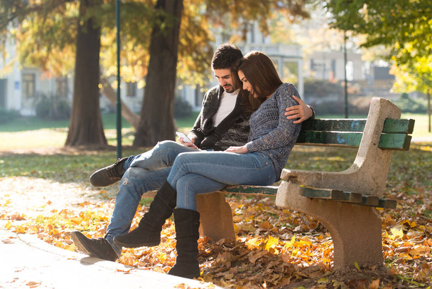 Young Couple Sitting on Bench in the Beautiful Autumn Day and Typing on the Phone In Park - Zdjęcie, obraz