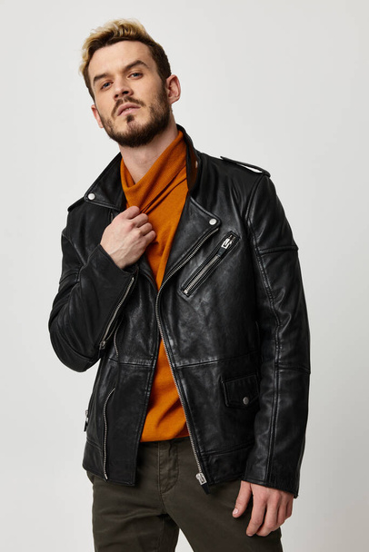 attractive young man in an orange sweater and in a leather jacket on a light background touches the collar - Foto, Imagen