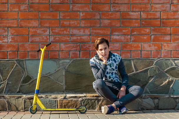 Un adolescent s'assied sur le trottoir à côté d'un scooter, et fronce les sourcils appuyé sur sa main. Mur de briques en arrière-plan. Le concept de l'adolescence et des émotions. - Photo, image