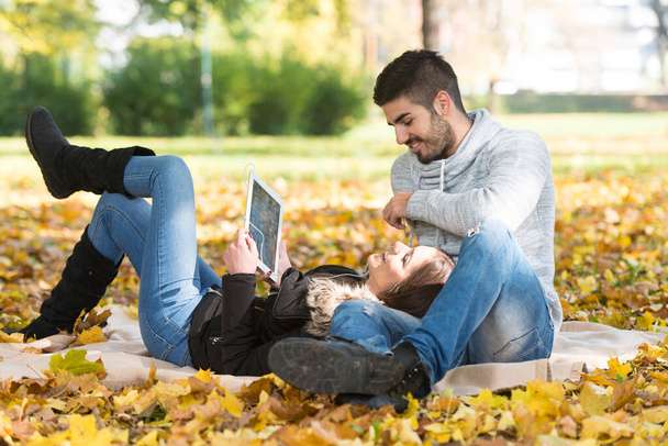 Young Couple Sitting on a Blanket in the Beautiful Autumn Day and Using Digital Tablet Listening on Headphones in Park - Foto, imagen