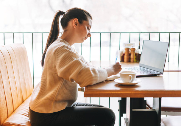 Atraktivní mladá brunetka usměvavá žena student na volné noze s šálkem čaje pomocí notebooku pracuje v kavárně - Fotografie, Obrázek