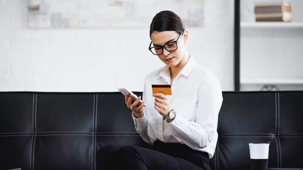 hübsche Geschäftsfrau sitzt mit Handy und Kreditkarte auf Sofa - Foto, Bild