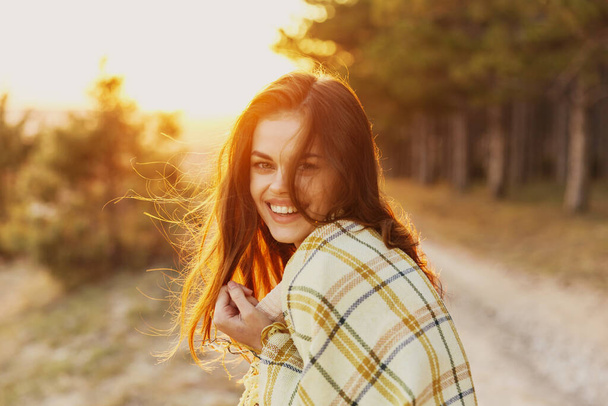 mujer con una manta en los hombros se ríe de la puesta de sol en el fondo y las coníferas en el lado - Foto, imagen