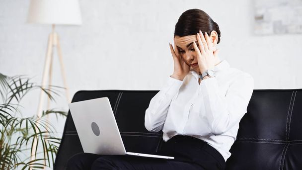 stanca donna d'affari che tocca la testa seduta sul divano con laptop - Foto, immagini
