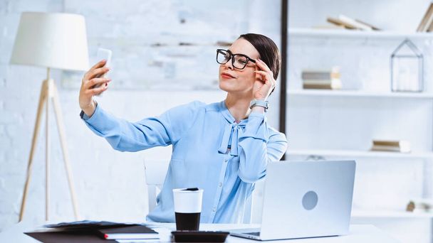 succesvolle zakenvrouw aanraken bril tijdens het nemen van selfie op de werkplek - Foto, afbeelding