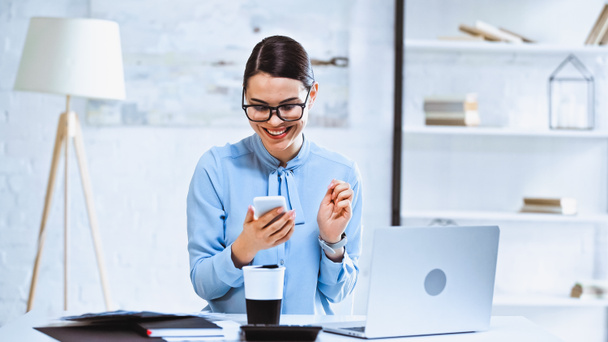 alegre mujer de negocios utilizando teléfono inteligente cerca de café para ir y portátil en el lugar de trabajo - Foto, Imagen