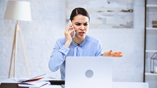 Wütende Geschäftsfrau spricht auf Smartphone, während sie im Büro auf Laptop schaut - Foto, Bild