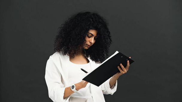 Кудрявая латиноамериканка, пишущая на планшете, изолированная на черном  - Фото, изображение