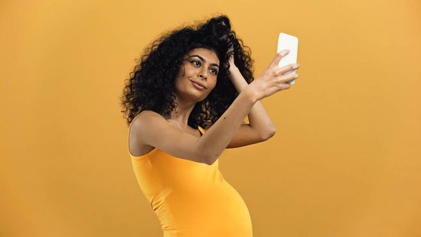 Těhotná hispánská žena užívající selfie s smartphonem izolované na žluté - Fotografie, Obrázek