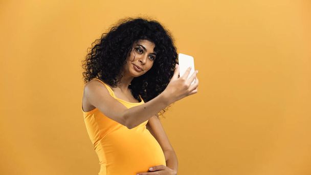 Έγκυος ισπανόφωνη γυναίκα που παίρνει selfie απομονωμένη σε κίτρινο - Φωτογραφία, εικόνα