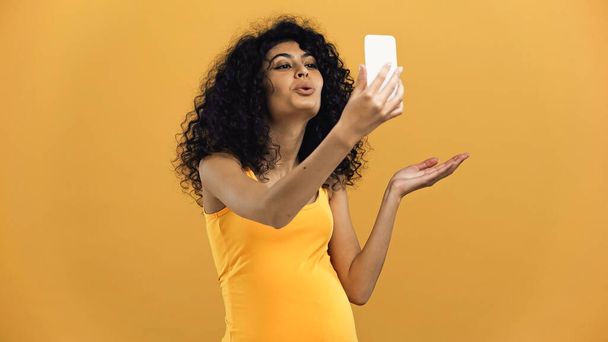 妊娠パニック女性吹い空気キス中にビデオコール上のスマートフォン隔離されましたオン黄色 - 写真・画像