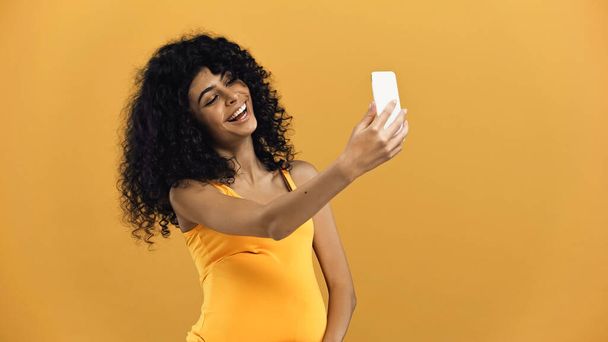 Веселая беременная латиноамериканка делает селфи на смартфоне, изолированном на желтом - Фото, изображение
