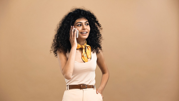 Mujer hispana joven con la mano en el bolsillo hablando en el teléfono celular aislado en beige  - Foto, Imagen