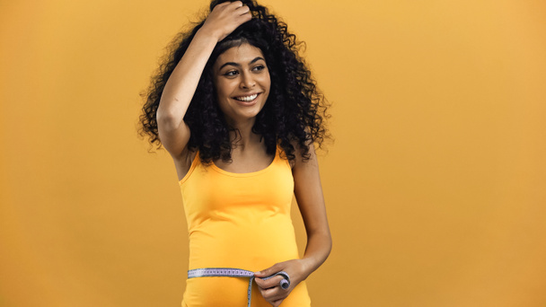 Happy pregnant hispanic woman holding tape measure isolated on yellow - Photo, Image