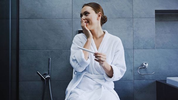 femme heureuse en peignoir tenant test de grossesse dans la salle de bain - Photo, image