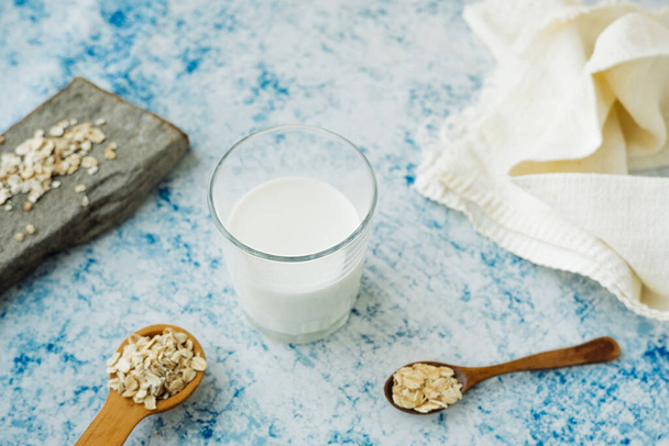 filiżanka mleka i drewniana łyżka z rdzeniem na niebieskim stole ozdobiona naturalnymi materiałami, prawdziwym kamieniem i bawełnianą serwetką. przepis na produkcję mleka owsianego w domu. zawartość żywności - Zdjęcie, obraz