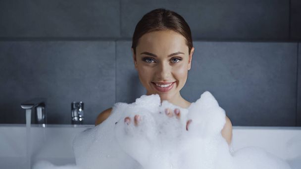 jeune femme souriante prenant un bain avec de la mousse dans une baignoire blanche  - Photo, image