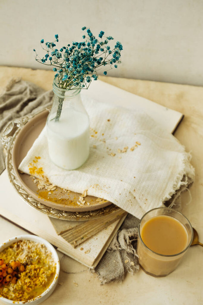 stylish simple monochrome beige table setting at home. gypsophila in glass bottle of milk on stack of old books, cup of coffee, plate of oatmeal, cotton napkins. creating comfort in interior - Photo, Image