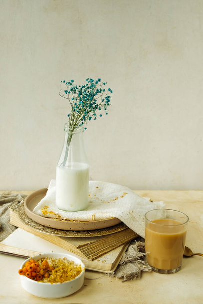 stijlvolle eenvoudige monochrome beige tafelschikking thuis. gypsophila in glazen fles melk op stapel oude boeken, kop koffie, bord havermout, katoenen servetten. comfort creëren in het interieur - Foto, afbeelding