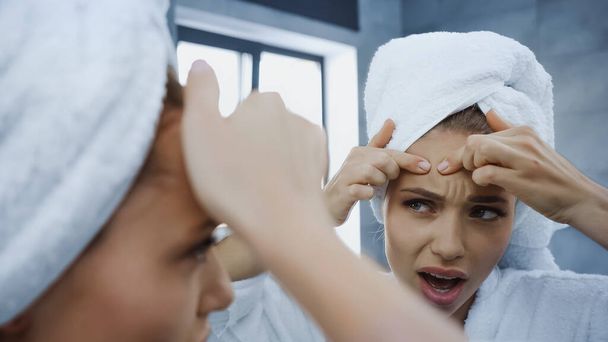 nespokojená mladá žena mačkání pupínek a pohled na zrcadlo v koupelně - Fotografie, Obrázek