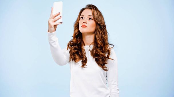 krullend jong vrouw het nemen van selfie geïsoleerd op blauw - Foto, afbeelding
