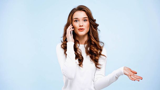 rizado mujer joven hablando en el teléfono inteligente aislado en azul - Foto, imagen