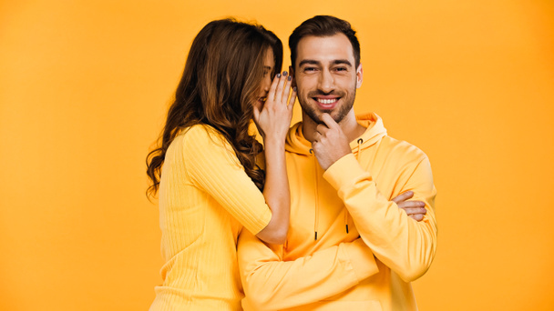 young woman whispering in ear of happy boyfriend isolated on yellow  - Photo, Image