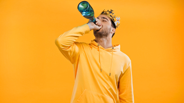 άνθρωπος στο στέμμα πίνοντας σαμπάνια από μπουκάλι απομονωμένο σε κίτρινο - Φωτογραφία, εικόνα